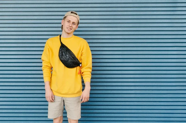 Bel ragazzo in elegante streetwear si erge sullo sfondo di un muro blu, sorride e guarda la fotocamera, indossa un cappello e un marsupio. Ritratto di un giovane uomo sorridente che indossa uno sfondo blu brillante — Foto Stock