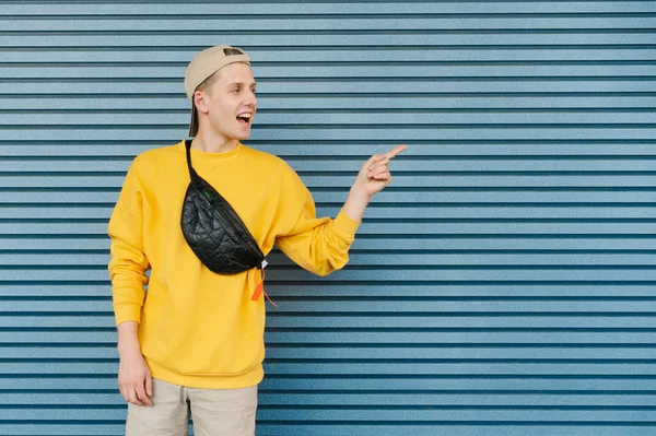 El joven en ropa de calle mira y muestra los dedos en la dirección de un lugar para la publicidad, con una gorra y una riñonera, aislados sobre un fondo de una pared azul. Copiar espacio —  Fotos de Stock