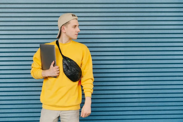 Studente con un computer portatile tra le mani e un marsupio intorno al collo in piedi sullo sfondo di una parete blu e guardando lateralmente allo spazio vuoto. Elegante freelance con un computer portatile posa — Foto Stock