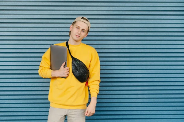 Beautiful guy in a cap and street stylish clothes stands on the background of a blue wall, holds a laptop in his hands, looks into the camera and smiles, wears a yellow sweatshirt and fanny pack.