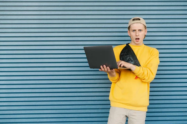 Portrait of a surprised boy stands with an open notebook on a blue background, looks into a camera with a shocking face, wears stylish streetwear, cap and a fanny pack. — Stock Photo, Image