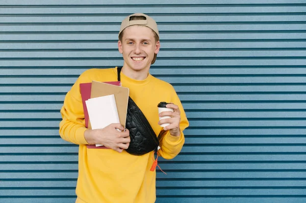 Улыбающийся студент в жёлтой толстовке и кепке стоит на фоне синей стены с книгами и блокнотами и чашкой кофе в руках, смотрит в камеру и улыбается. Копирование пространства — стоковое фото