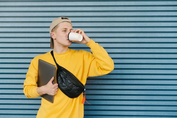 Портрет стильного парня в уличной одежде, кепках и поясной сумке, стоящего на синем фоне с ноутбуком в руке и пьющего кофе из бумажной чашки, смотрящего боком — стоковое фото