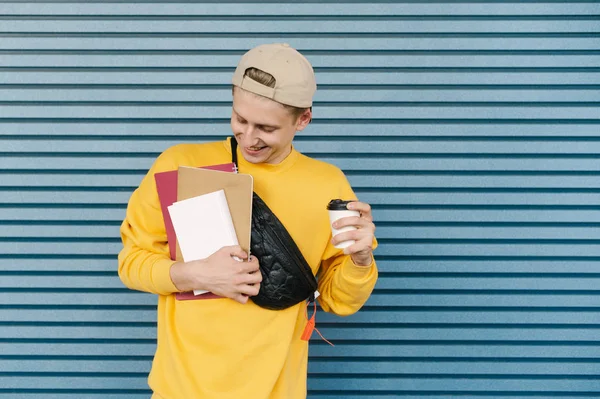 Estudiante con gorra y sudadera amarilla se levanta sobre el fondo de una pared azul con libros, cuadernos y una taza de café en las manos, sonríe y mira hacia abajo. Aislado contra el fondo de la pared — Foto de Stock