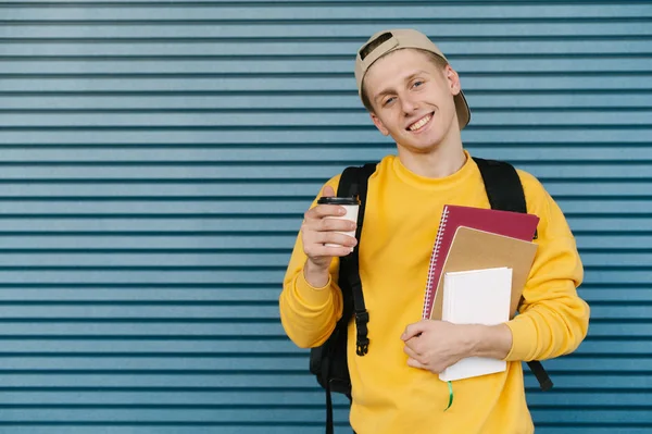 一个穿着鲜艳衣服的快乐的年轻学生站在蓝色墙壁的后面对着相机摆姿势，拿着一杯咖啡和一本书，看着相机笑了。. — 图库照片