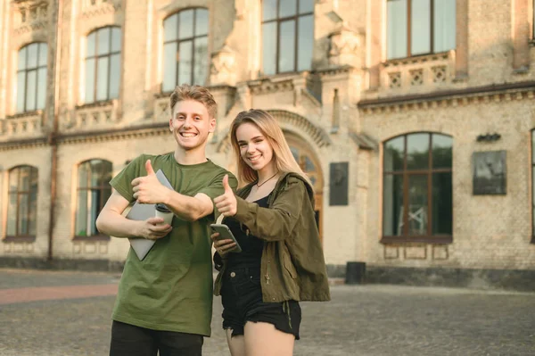 Счастливая пара успешных студентов улыбается, показывая большие пальцы на камеру возле кампуса колледжа. Два веселых студента поднимают большие пальцы у универа, держа в руках ноутбук, кофе и смартфон . — стоковое фото
