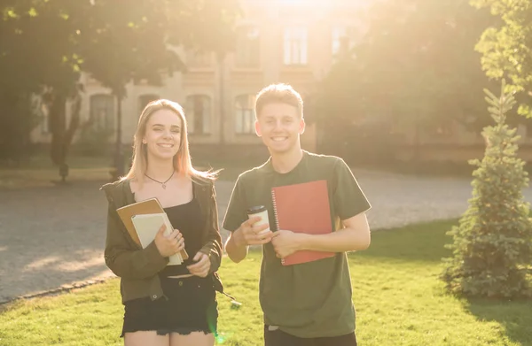 Dos estudiantes emocionados con exámenes aprobados cerca de la universidad sosteniendo los cuadernos y llevar la taza de café. Un par de estudiantes en el parque cerca de la universidad sonriendo y sintiéndose descuidados . Fotos De Stock