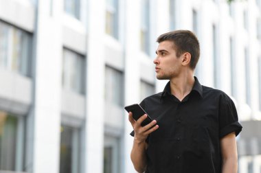 Siyah tişörtlü çekici genç bir ofis çalışanı elinde akıllı telefonuyla açık renkli bir arka planda duruyor ve boş bir alana bakıyor. Şık bir ofis çalışanının portresi.