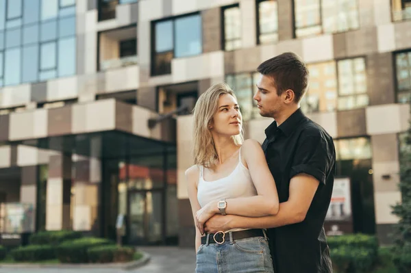 这对年轻漂亮的夫妇在一座现代建筑的背景下拥抱在大街上 女孩把目光移开了 在一个温暖的夏日里 男人拥抱着女孩散步 她很开心 爱情故事 — 图库照片