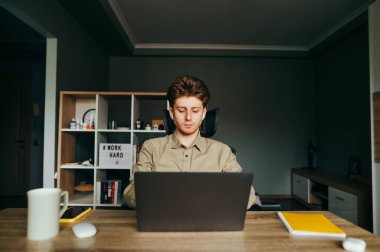 Kablosuz kulaklıklı, iş yerinde dizüstü bilgisayarla çalışan, ekrana odaklanan ciddi genç bir iş adamı. Uzaktan kumandayla evde çalışmakla meşgul. Karantina.