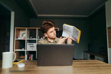 Gömlekli ve kulaklıklı bir çalışan, meslektaşları ve patronuyla çevrimiçi bir toplantıda web kamerasına bir not defteri gösteriyor. Adam evde karantinada ve video görüşmeleri üzerinde çalışıyor..