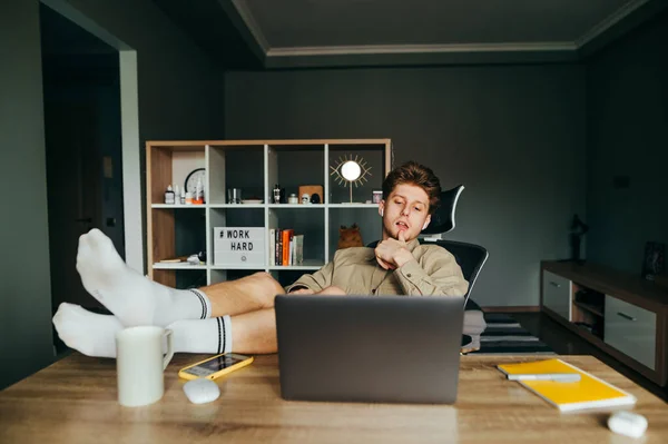 Relaxed freelancer lies in a chair at work at home with his legs raised on the table and uses a laptop, wears a shirt and home shorts on his feet. Remote work in quarantine concept.