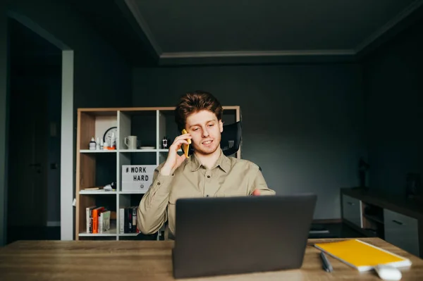 A positive business man works remotely from home, sits on a work chair at home and talks on the phone against the backdrop of a cozy bedroom. The freelancer works at home on a laptop and calls.