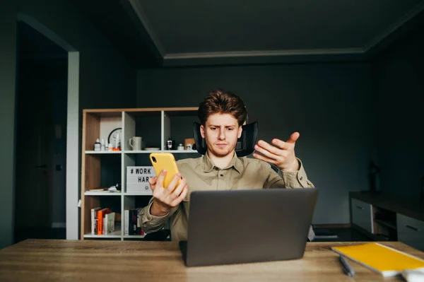 Dissatisfied employee in a shirt sits at home on the remote with a laptop and smartphone in hand, dissatisfied looking at the computer screen and gesturing. Dissatisfied freelancer works at home.