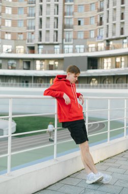 Kırmızı kapüşonlu atletik genç adam Velodrome 'un arka planında duruyor ve koşudan sonra dinleniyor. Eğitimdeki yorgun adam arka planda yanıklar var. Dikey.