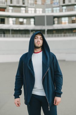 Mavi mantolu sakallı bir adamın portresi kentsel bir arka planda duruyor ve ciddi bir yüzle kameraya poz veriyor..