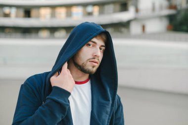 Kapüşonlu başlıklı sakallı yakışıklı adamın yakın portresi. Şehir arka planında ciddi bir yüzü olan kameraya bakıyor..