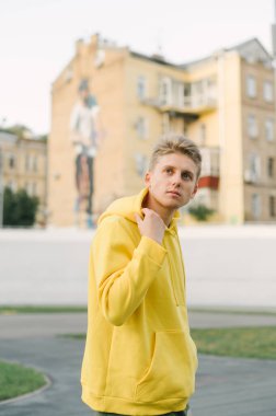 Sarı kapüşonlu yakışıklı bir gencin portresi spor sahasında duruyor ve kentsel bir arka planda kameraya poz veriyor. Dikey.