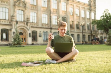 Üniversitenin yanındaki çimlerde oturan gülümseyen genç adam elinde bir fincan kahveyle dizüstü bilgisayar kullanıyor. Üniversite kampüsündeki çimlerin üzerinde oturmuş pencereye bakıyor ve gülümsüyor..
