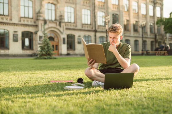 Thoughtful young man in casual clothes is sitting with a laptop on the grass on a college campus and reading a notebook with a worried face. Student learns a task during a break at university.
