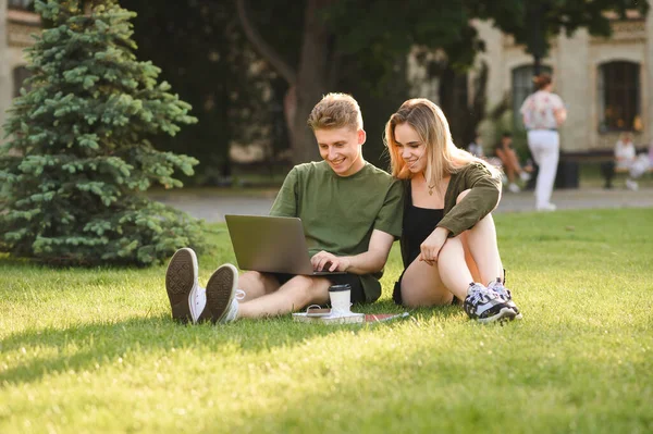 一对快乐的学生坐在大学校园草坪上 一边用笔记本电脑 一边看着屏幕 一边笑着 在公园休息的时候 带着笔记本电脑学习的女孩和年轻人在笑 学生闲暇时间 — 图库照片