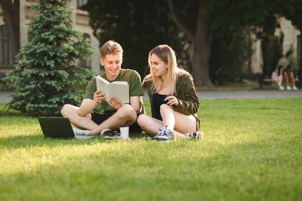 大学の公園の芝生の上に本やノートパソコンを持って座って 笑顔で顔を勉強し 話している前向きな男と女の子 キャンパスで草の上で一緒に過ごす幸せな学生の美しいカップル — ストック写真