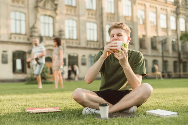 Portrait Hungry Young Man Sitting Grass Backdrop University Building Eating — Stock Photo, Image