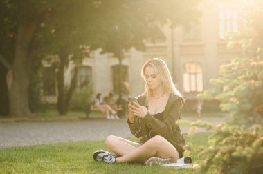 Çekici kız gün batımında kampüste kitaplarla oturuyor, akıllı telefon kullanıyor ve kulaklıkla müzik dinliyor. Güzel kız öğrenci üniversite arka planında akıllı telefon kullanıyor..