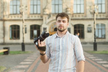 Yakışıklı genç fotoğrafçı elinde kamerayla mimari arka planda kameraya bakıyor ve gülümsüyor. Elinde profesyonel kamera olan yakışıklı sakallı fotoğrafçının sokak portresi..