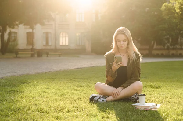 カジュアルな服を着たかわいい女の子は 本やコーヒーを片手に大学公園の芝生の上に座り 真剣な顔をしたスマートフォンを使用しています スマート魅力的な女の子は クラスの後に休憩のためにスマートフォンを使用します — ストック写真