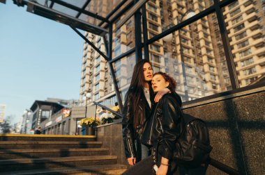 Şehir arka planında merdivenlerde dikilen iki kızın sokak fotoğrafı, kameraya bakıp poz verirken, şık sokak kıyafetleri ve sırt çantası giyerken. Arka planda sarılan iki manken. Boşluğu kopyala