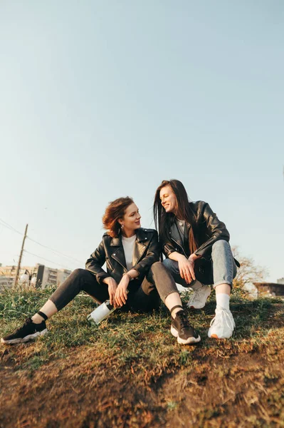 通りの服で2人の幸せな女の子が草の上に座っている ワインのボトルを保持し お互いを見て 空と都市の背景に笑みを浮かべている ガールフレンドは散歩を楽しんでいます — ストック写真