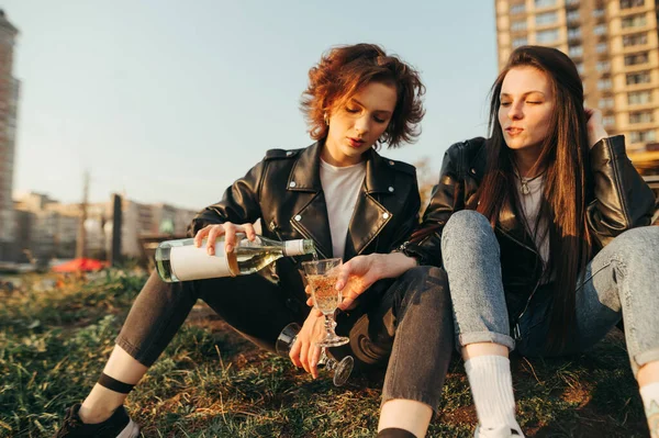 两个穿着时髦衣服的漂亮姑娘坐在草地上 背对着城市景观 把葡萄酒倒入杯子里 快乐的女孩在城市里散步时喝酒 坐在草地上放松 — 图库照片