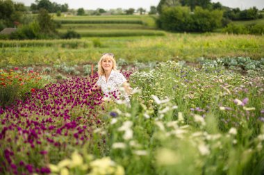 Çekici sarışın kadın bir kulübede bir yatakta oturmuş çiçeklerin arasında kameraya poz veriyor. Hobi olarak çiçek yetiştirmek..