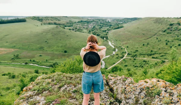 Hipster woman stands with her back to a rock and looks at a beautiful green landscape with a river and a valley. The tourist stands on a hill and admires the landscape. Domestic tourism