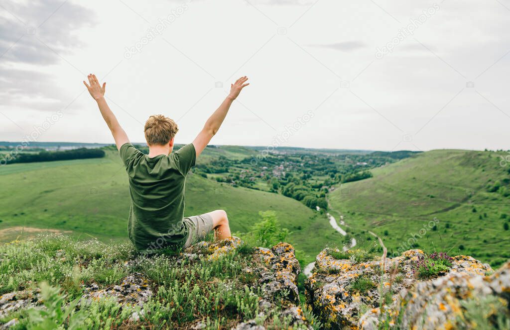 Joyful young male tourist sits with his back on a rock cliff and looks at the beautiful green landscape, raised his hands up. Male hiker feels freedom sitting on top of a hill.