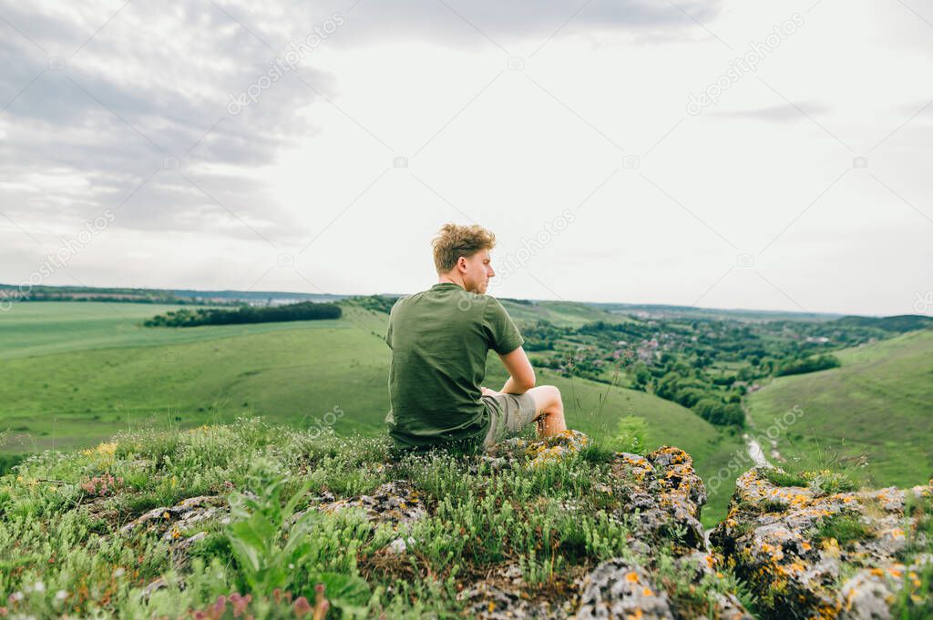 A young man in casual clothes sits on a cliff on a rock and looks at the beautiful Ukrainian landscape from a height. A tourist is resting on top of a mountain in Ukraine. Tovtry.