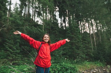 Çekici ve mutlu turist kız ellerini havaya kaldırıyor ve köknar ormanında neşeli hissediyor. Olumlu ve neşeli genç kadın ormanda heyecanlı, eller havada, geniş bir gülümseme.