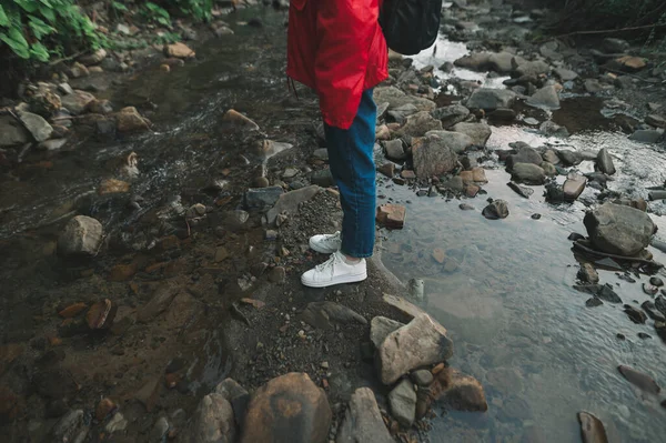 山の川の真ん中に石の上に立って赤いレインコートの少女の足の写真 渓流の水の近くに立つ白いスニーカーの女性観光客の夜の写真 — ストック写真