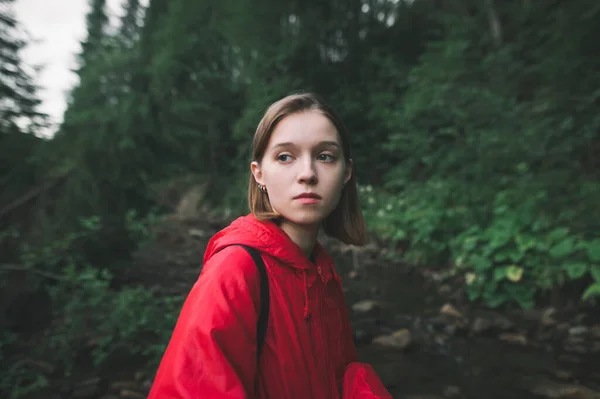 Close up portrait of a female hiker in a red jacket is calm and relaxed in the fir forest. Young tourist girl feeling upset and disappointed in the mountain woods near the river.