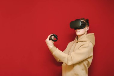 Kırmızı arka planda kontrolörleri olan parlak kapüşonlu genç bir adamın fotoğrafı ve video kaskla oyunlar oynayıp, gözlerini kaçırması. VR oyun kaskı konsepti. Boşluğu kopyala