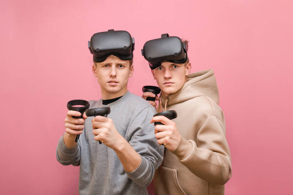 Two young men stand on a pink background with VR helmets on their heads and controllers in their hands, looking intently into the camera on a pink background. Friends play VR games on console.