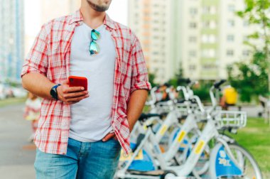 Günlük kıyafetli bir adam elinde akıllı bir telefonla bisiklet kiralama parkının arka planına dayanıyor ve bisiklet paylaşım programının keyfini çıkarıyor. Bisiklet kiralama çevrimiçi