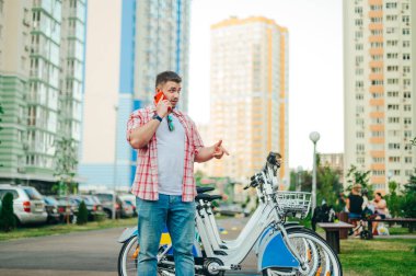 Parlak günlük kıyafetli bir adam arka fonda duruyor. Kiralık bisikletleri var ve operasyon telefonuyla yüzünde bir gülümsemeyle iletişim kuruyor. Sokakta bisiklet kiralama. Bisiklet paylaşımı