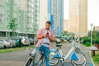 Sakallı ve günlük kıyafetleri olan bir adam bisikletini paylaştı ve yüksek binaları olan bir avlunun arka planına karşı akıllı telefon kullandı. Adam bisiklet kiralamak için mobil uygulama kullanıyor.