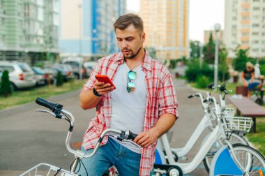 Gezici bir uygulama üzerinden çevrimiçi yürüyüş için bisikleti paylaşan turist, bir dizi bisiklet ve manzaraya karşı ciddi bir yüze sahip bir akıllı telefon kullanıyor.