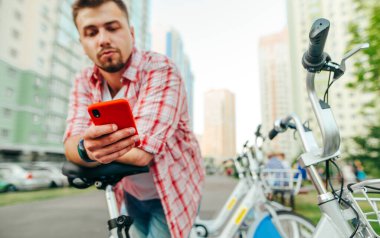 Bir erkek turist, bisikletle sırada bekliyor ve akıllı telefondan paylaşmak için mobil bir uygulama kullanıyor. İnsanoğlunun elindeki akıllı telefona odaklan. Bisiklet paylaşımı