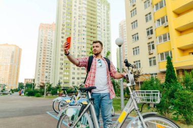 Pozitif genç adam sokakta kiralık bir bisikletle duruyor ve şehrin arka planına karşı selfie çekiyor. Akşamları bisiklet süren ve akıllı telefondan fotoğraf çeken bir adam.