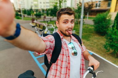 Bisikletli komik bir adam sokakta selfie çeker, akıllı telefonun kamerasına bakar ve komik bir yüz ifadesi takınır. Neşeli turist bisiklet sürerken fotoğraf çekiyor.