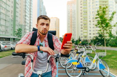 Sakallı yakışıklı bir adam şehrin arka planında duruyor ve paylaşmak için bisiklet serisi, akıllı telefon kullanıyor ve ciddi bir yüzle kameraya bakıyor. Sokakta bisiklet sürüyor.